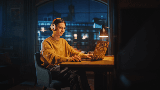 Person mit Kopfhörern arbeitet abends am Laptop, warmes Schreibtischlicht und Stadtfenster im Hintergrund.