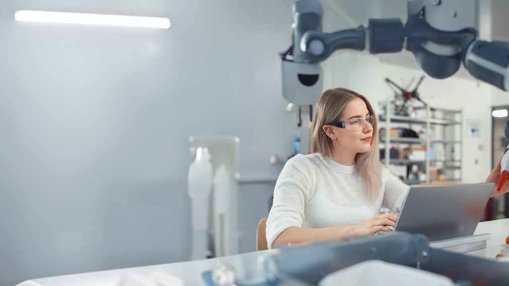 Eine Frau mit Schutzbrille arbeitet neben einem Roboterarm an einem Laptop in einem modernen Techniklabor.